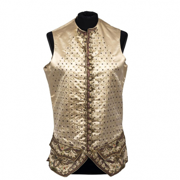 Chupa en raso de seda de color marfil, cuello a la caja y sin mangas. Cerrada en el delantero con botones de seda decorados con cordoncillo dorado y bordado floral. con hilos y aplicaciones de plata y oro. En la parte inferior, bolsillos con cartera en los faldones.