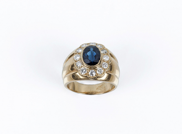 Sortija ancha en sólida montura gallonada de oro amarillo, con bello zafiro azul oval central, en chatón, y orla de limpios y blancos diamantes talla brillante.
