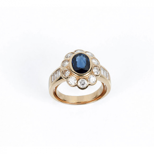 Sortija en s&oacute;lida montura de oro amarillo 18 K con zafiro fino azul, oval, en chat&oacute;n, y orla en fila de limpios y blancos diamantes, 