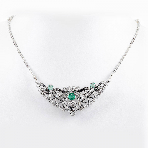 Collar gargantilla en oro blanco con gran centro formado por alfiler desmontable en forma de doble palmeta vegetal y lazada, con tres centros de esmeraldas talla brillante y cuajado de blancos diamantes talla rosa y talla simple