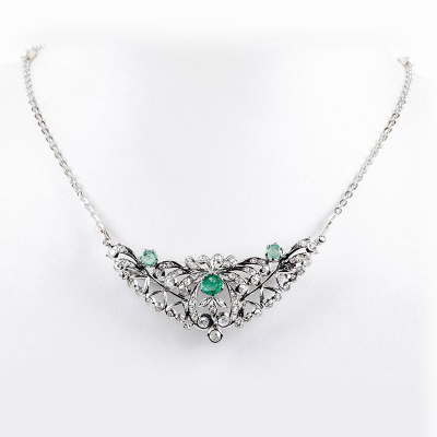 Collar gargantilla en oro blanco con gran centro formado por alfiler desmontable en forma de doble palmeta vegetal y lazada, con tres centros de esmeraldas talla brillante y cuajado de blancos diamantes talla rosa y talla simple