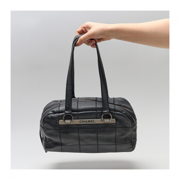 CHANEL. Bolso modelo «Boston Choco Bar» en cuero negro con trama de costuras y placa decorativa logotipada en metal antracita.